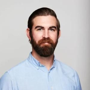 A bearded person with short, slicked-back hair wearing a light blue collared shirt.