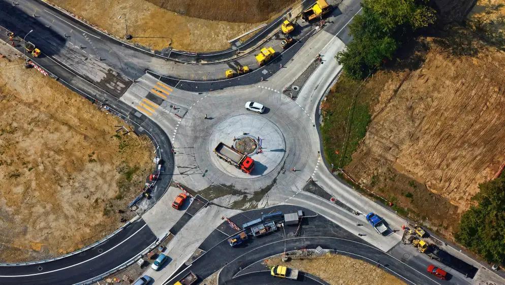 Aerial view of a multi-lane roundabout under construction, with various construction vehicles and machinery surrounding it. The landscape includes dirt and grassy areas, as well as partially finished roads with vehicles navigating the roundabout.