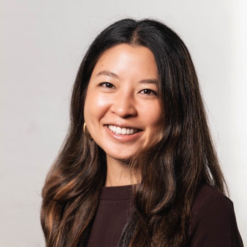A person with long dark hair smiles warmly, wearing a dark top, against a plain white background.