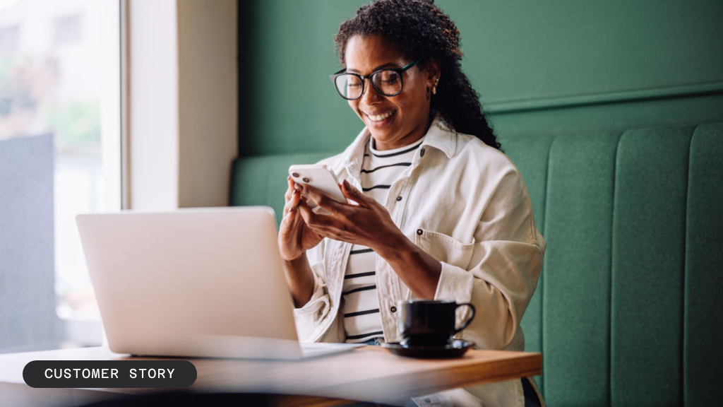Smiling Black woman wearing glasses, looking at her smartphone with a laptop and coffee on the table.