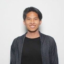 A person with short, dark hair smiles at the camera, wearing a black shirt and a dark, textured jacket against a plain, light background.