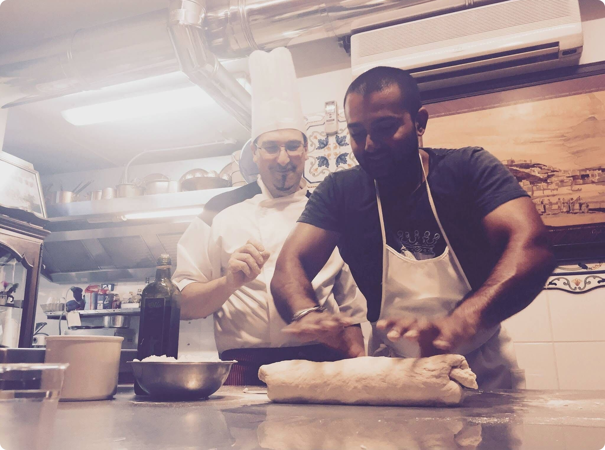 Two men in a kitchen, one wearing a chef's hat and the other an apron, are preparing dough on a countertop cluttered with cooking ingredients and utensils.