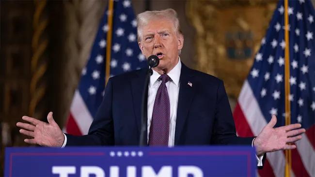 A man in a suit and tie speaks at a podium with flags in the background.