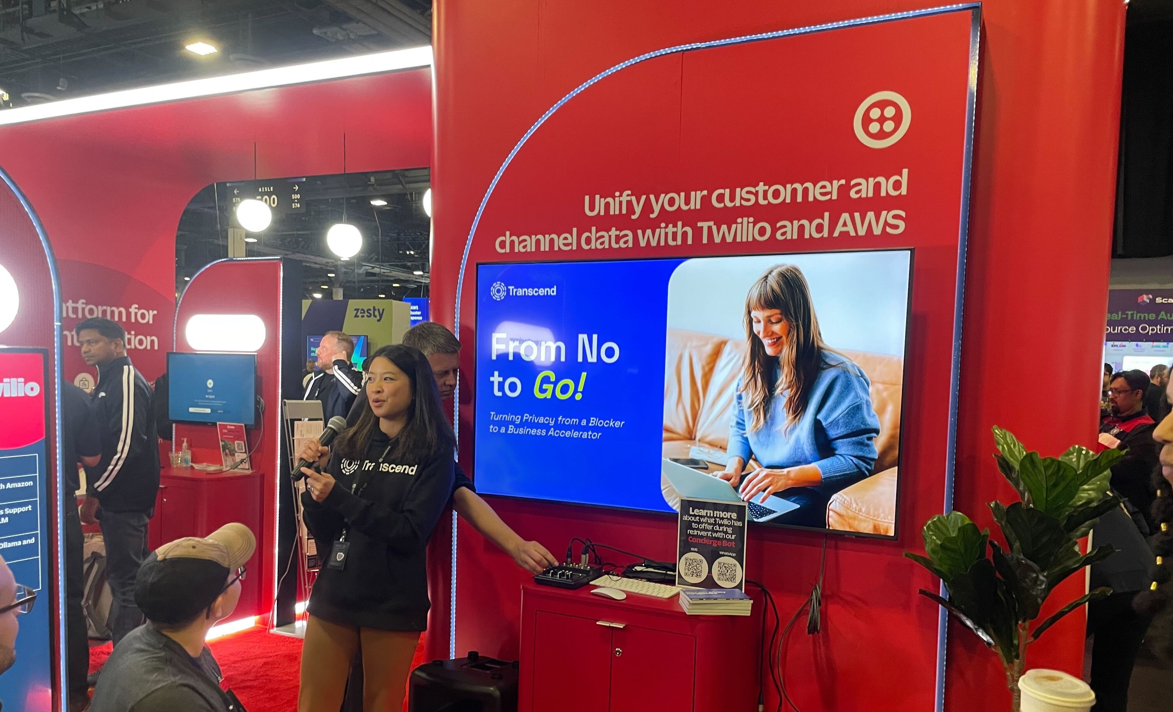 A booth at a tech event features a large red display with the Twilio and AWS logos. A screen shows a woman using a laptop with the text "From No to Go!" while a representative engages attendees nearby.
