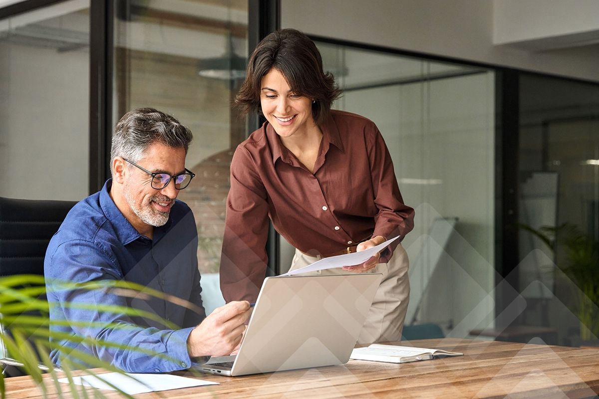 Two smiling colleagues collaborating on a laptop at an office desk.