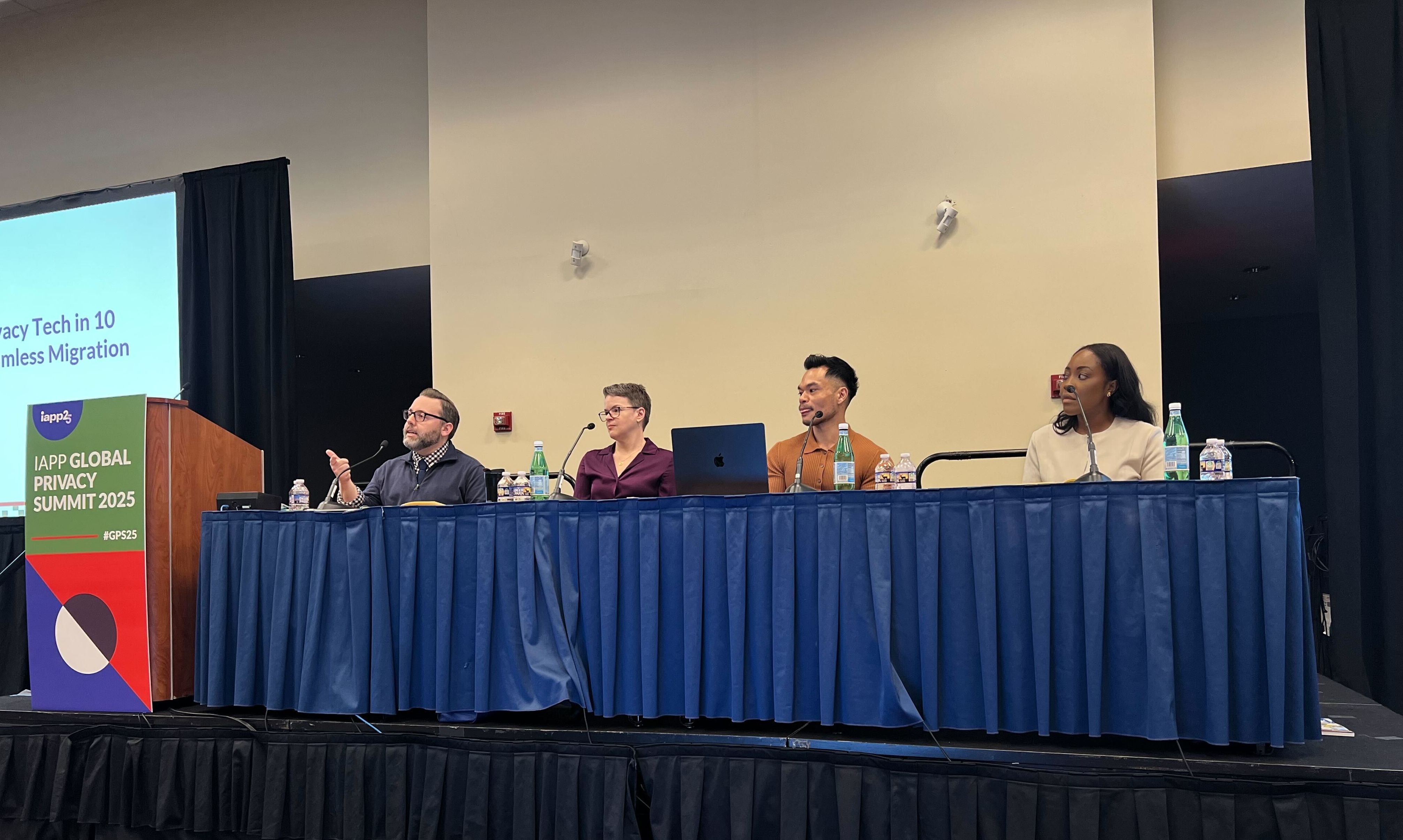 A panel of four people is sitting at a long table on a stage, each with microphones and laptops. They are participating in a discussion at the IAPP Global Privacy Summit 2025.
