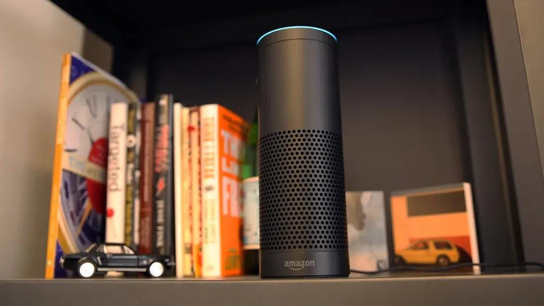 A black Amazon Echo speaker stands on a shelf in front of several books, with a small toy car placed beside it.