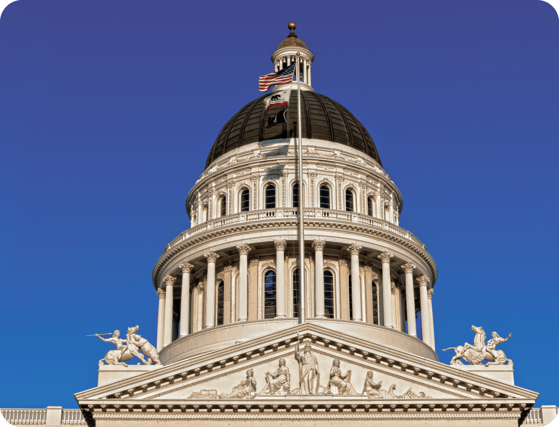 The dome of a classical government building with an American flag and intricate sculptures, set against a clear blue sky.