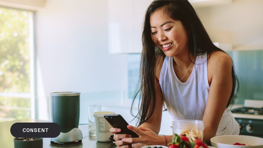 Smiling Asian woman looking at her phone in a kitchen with a blender and fruit.