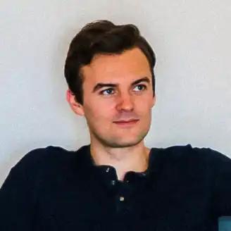 A man with short brown hair, wearing a dark shirt, looking slightly to the side with a neutral expression against a plain background.