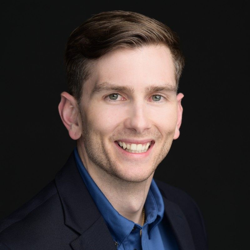 A smiling man with short brown hair, wearing a blue shirt and a dark blazer, against a black background.