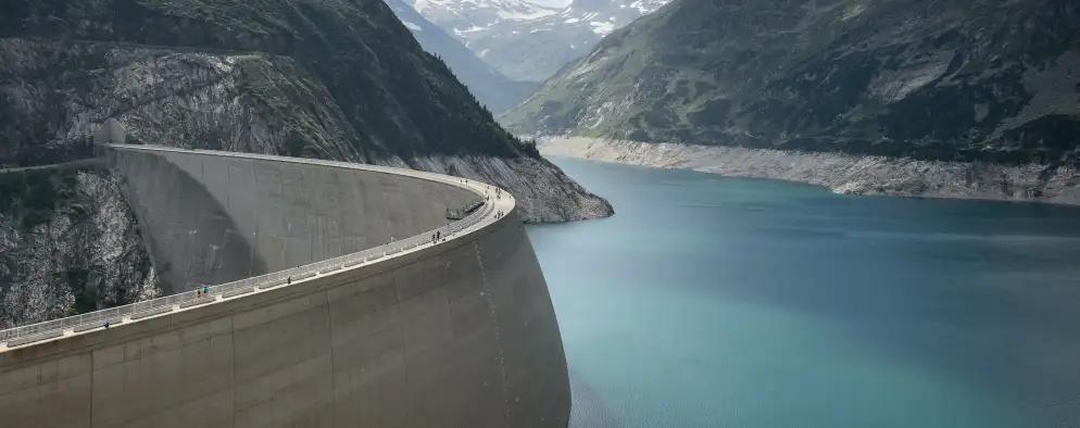 A large concrete dam curves along the edge of a deep turquoise reservoir, surrounded by steep, rocky mountains under a partly cloudy sky.