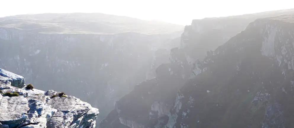 A rocky cliff edge overlooking a misty canyon with rugged terrain and a hazy sky in the background.