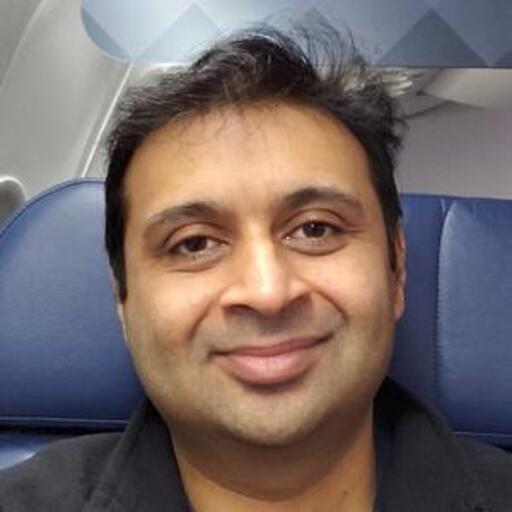 A person with short dark hair and a slight smile is seated in an airplane, with a blue seat and a window visible in the background.