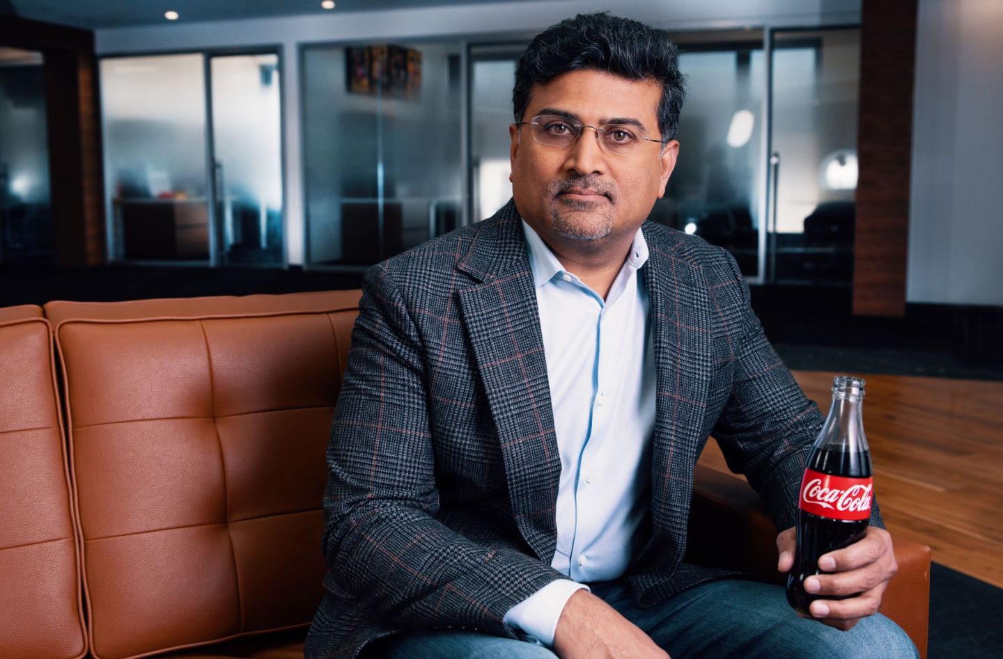 A man in a checkered blazer and glasses sits on a brown leather couch holding a bottle of Coca-Cola, with a modern office setting in the background.