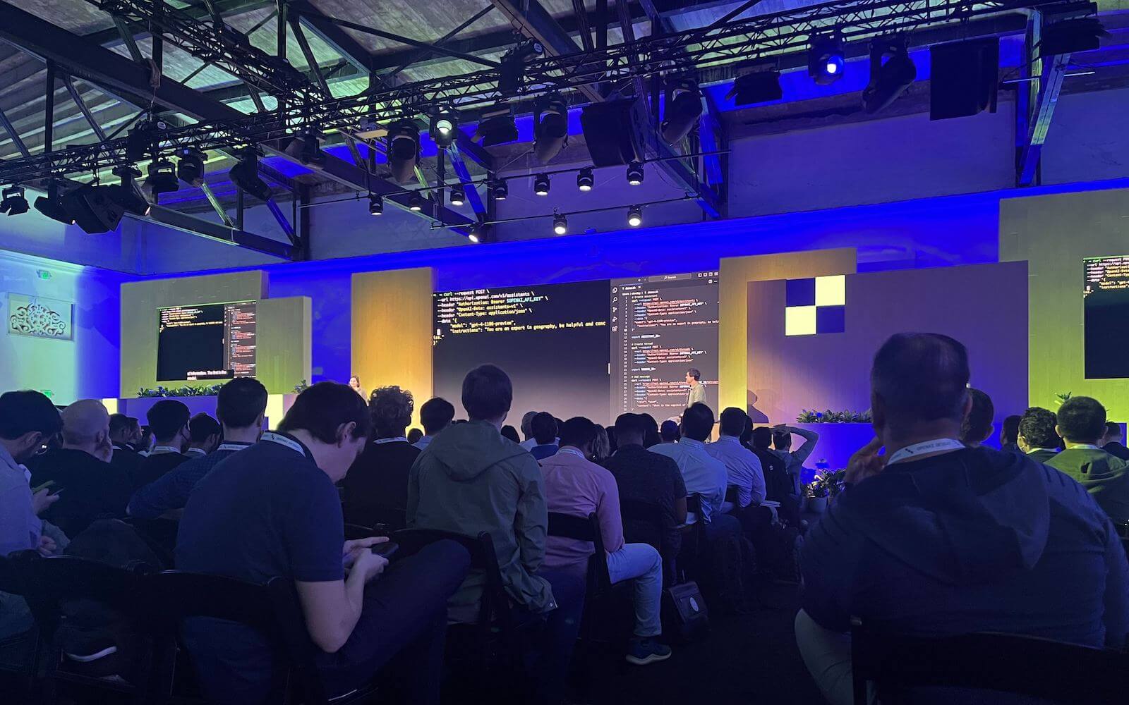 Speaker panel at OpenAI dev day showing the view of the stage from the crowd with code on screen