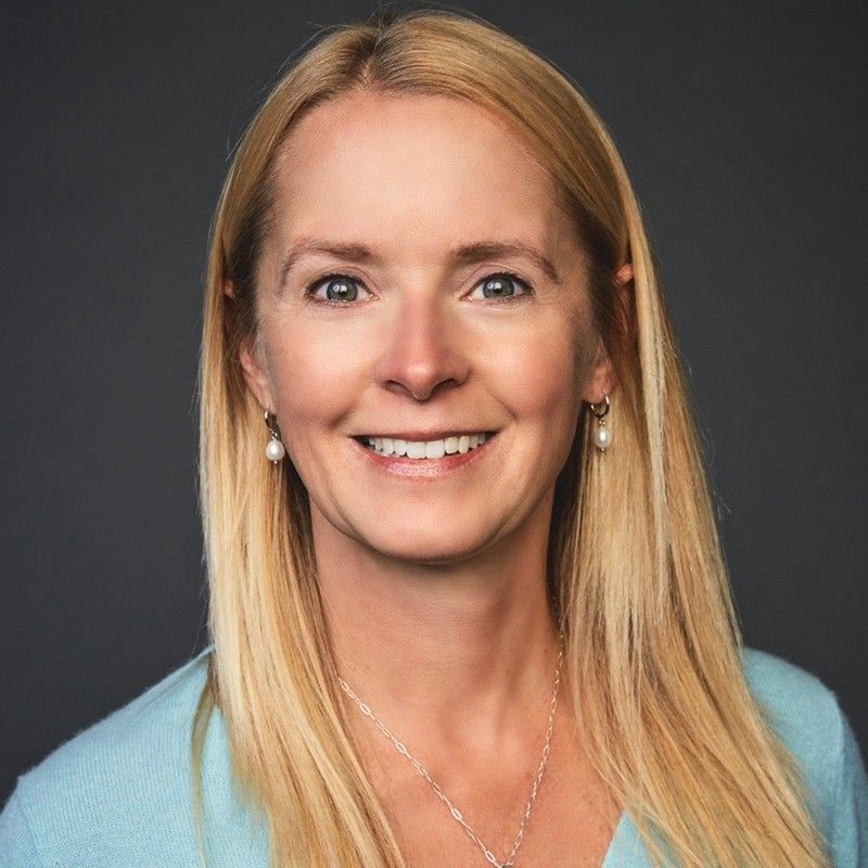 A smiling woman with long blonde hair, wearing pearl earrings, a light blue top, and a silver necklace, against a dark background.