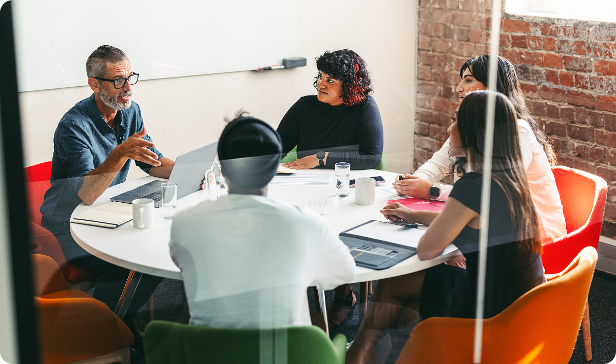 Group of professionals sitting in a conference room at a round table listening to a man with a laptop speak