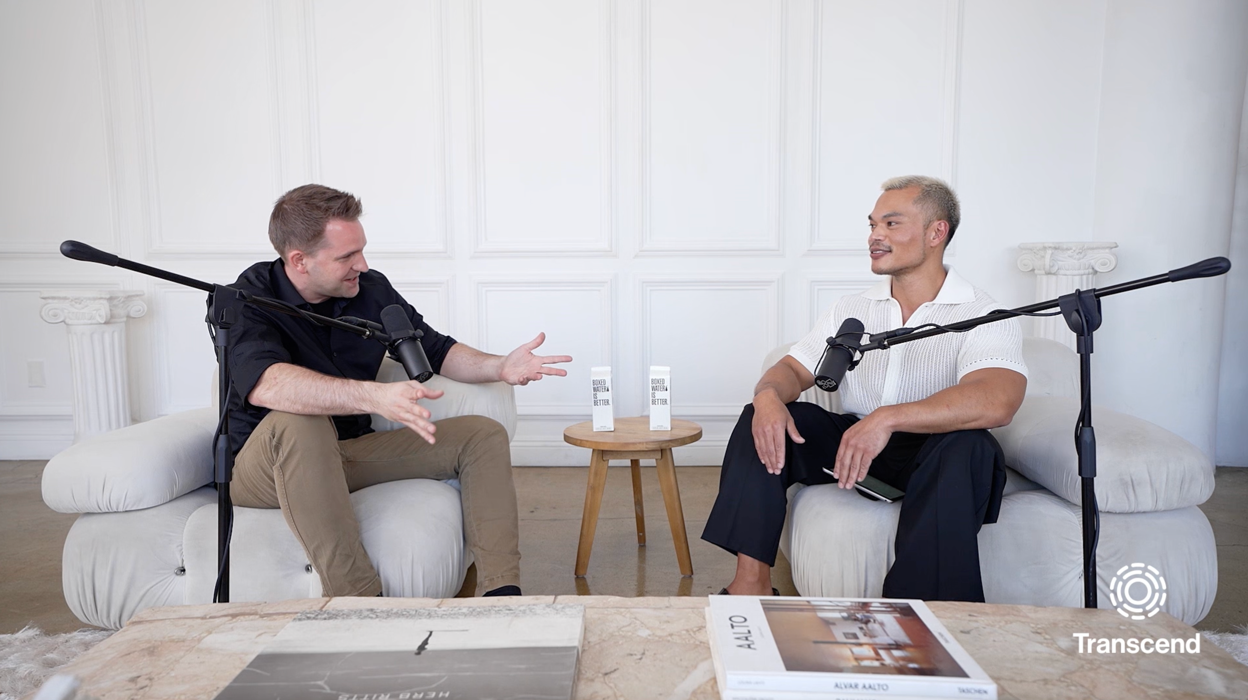 Two people sit on comfortable chairs in a modern white room with microphones in front of them, engaged in conversation. A small table with products is placed between them, and books are on a nearby table.