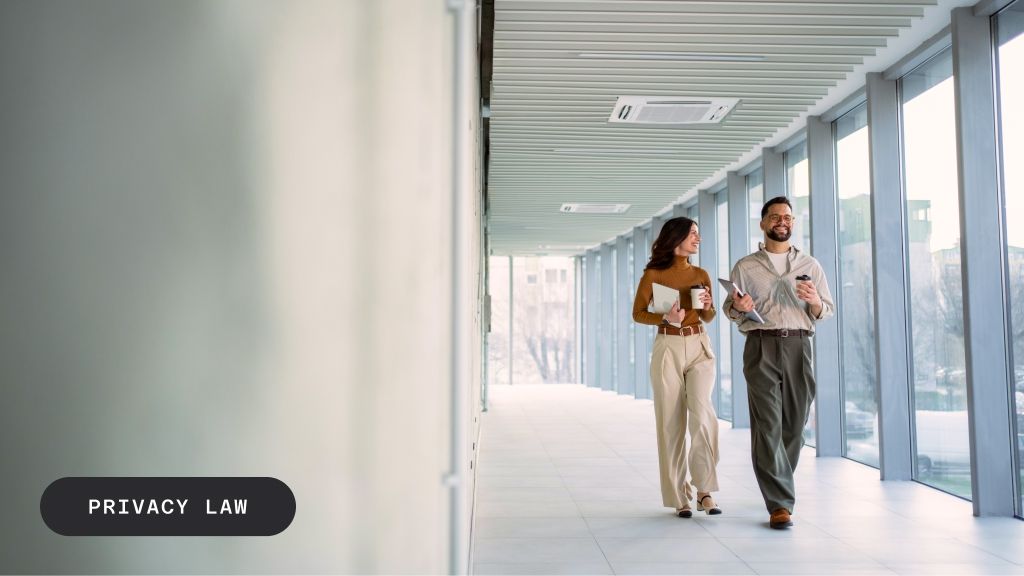 Two smiling professionals walk down a bright office hallway with large windows. Text overlay: "PRIVACY LAW".