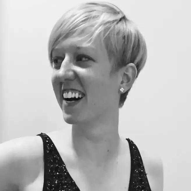 A smiling person with short blonde hair, wearing a sleeveless, glittery top, and small square earrings, looking slightly to the side.