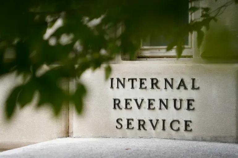 The entrance to a building displaying the words "Internal Revenue Service" in bold letters on the wall, partially obscured by tree branches.
