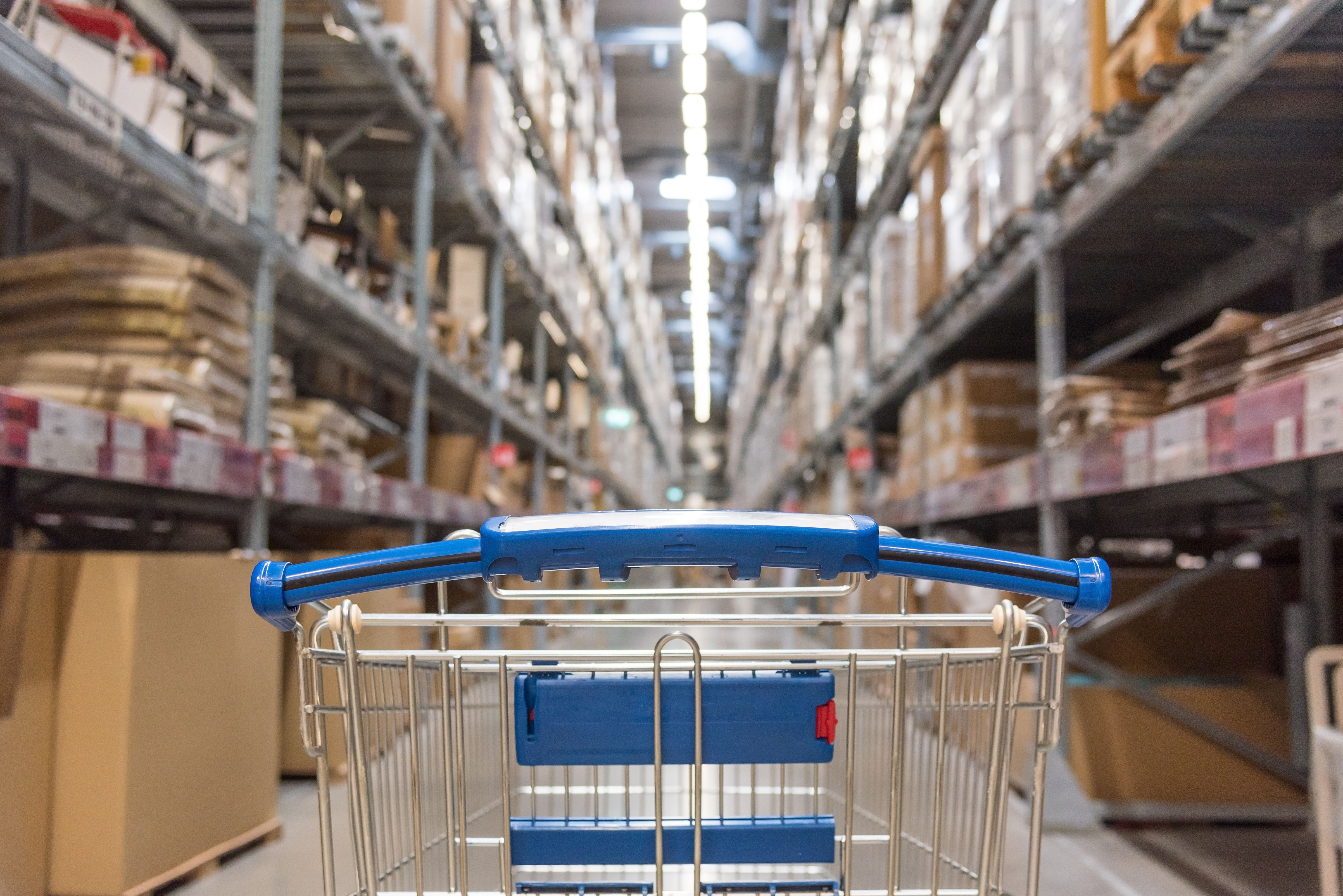 A shopping cart in a long warehouse aisle with shelves of boxes.
