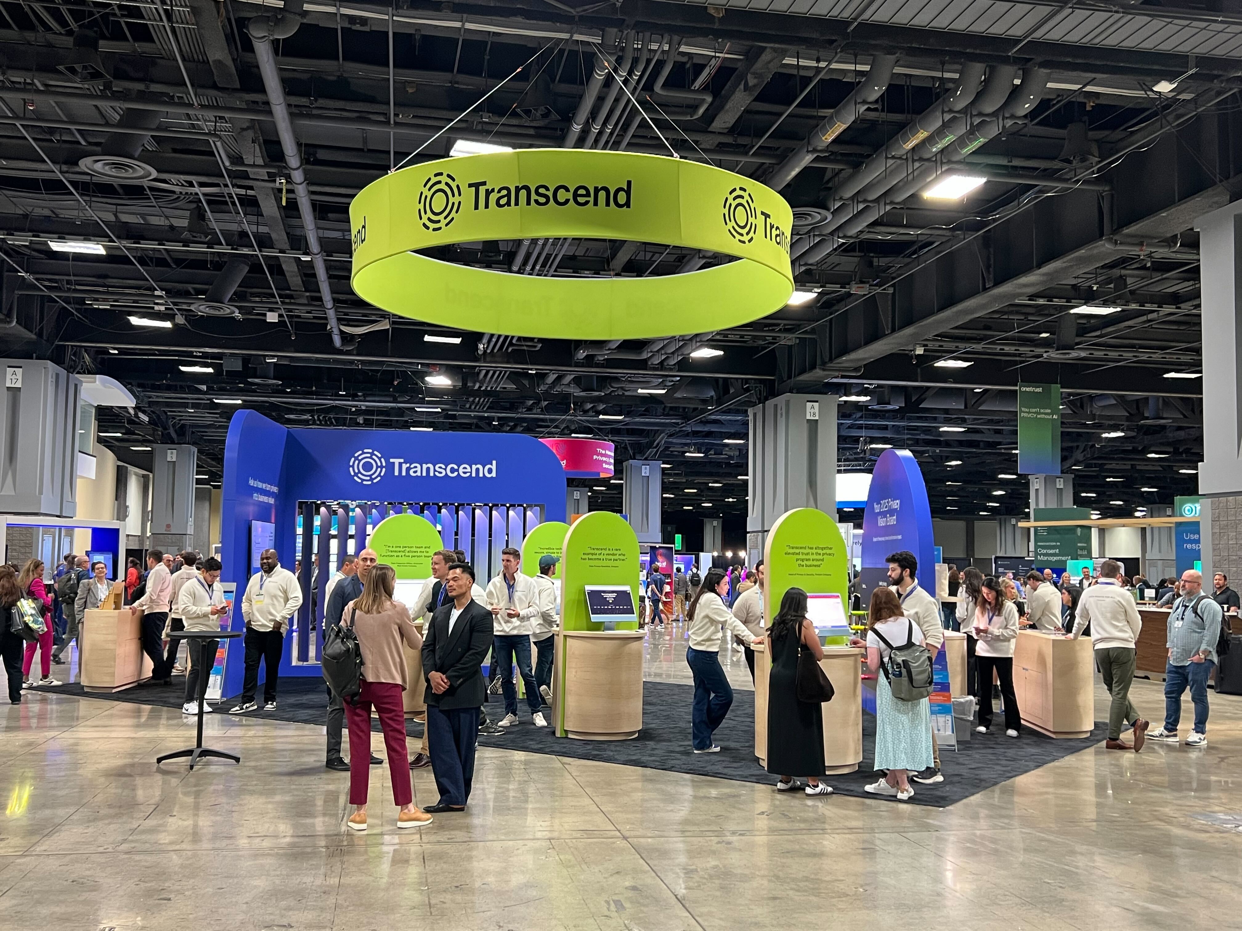 A bustling convention center features a Transcend booth with staff interacting with attendees. Bright green and blue colors dominate the display, while a circular banner with the Transcend logo hangs overhead.