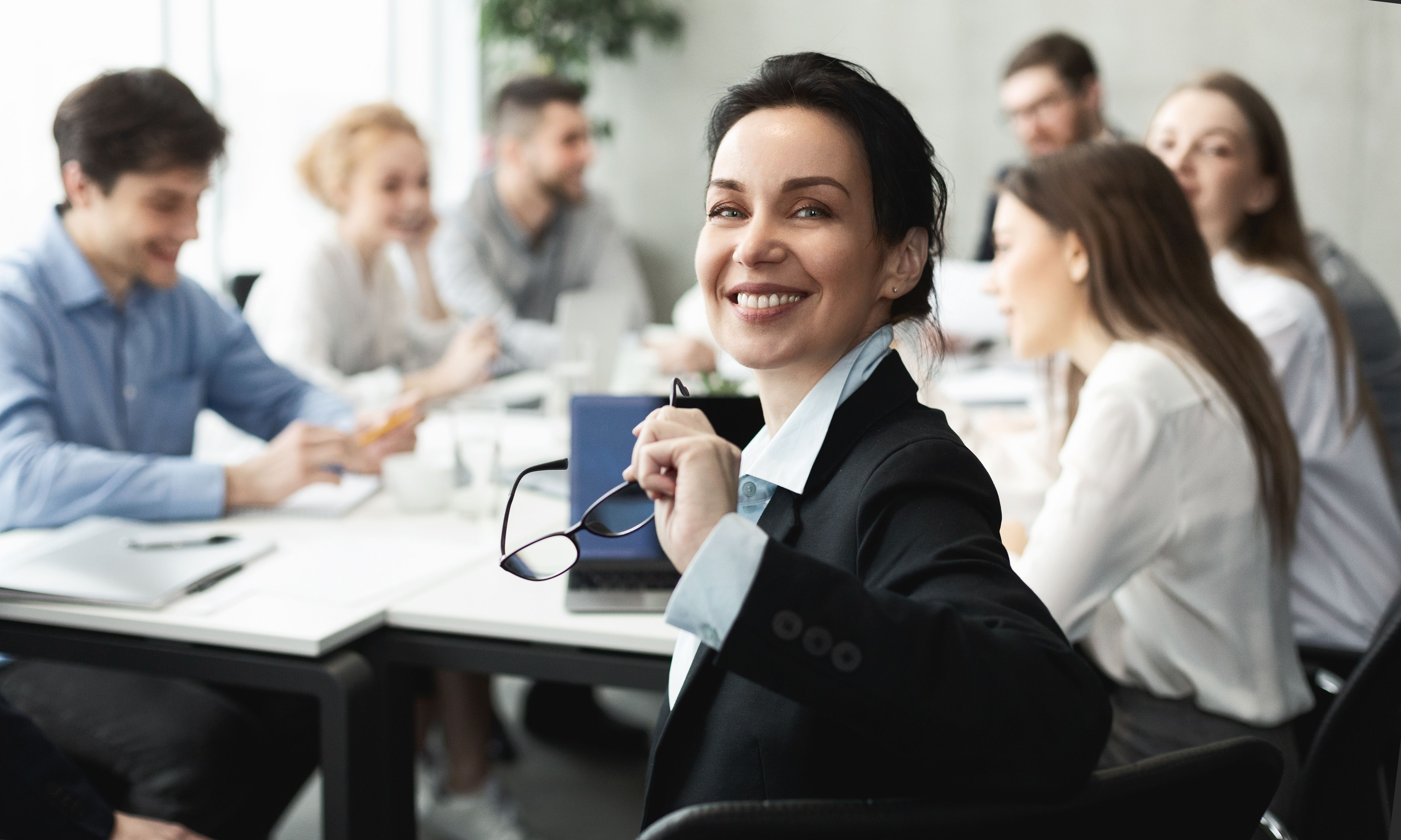 Smiling businesswoman holding glasses, with blurred colleagues in a meeting.