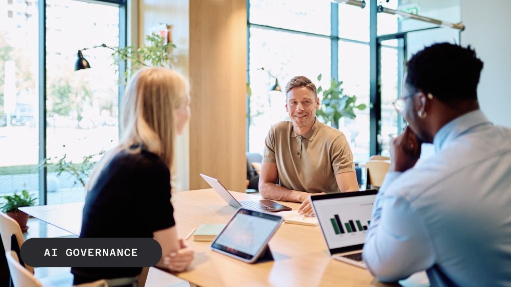 Three people meet in a modern office, with a man smiling towards the viewer and a laptop displaying a bar graph. Text: AI GOVERNANCE.
