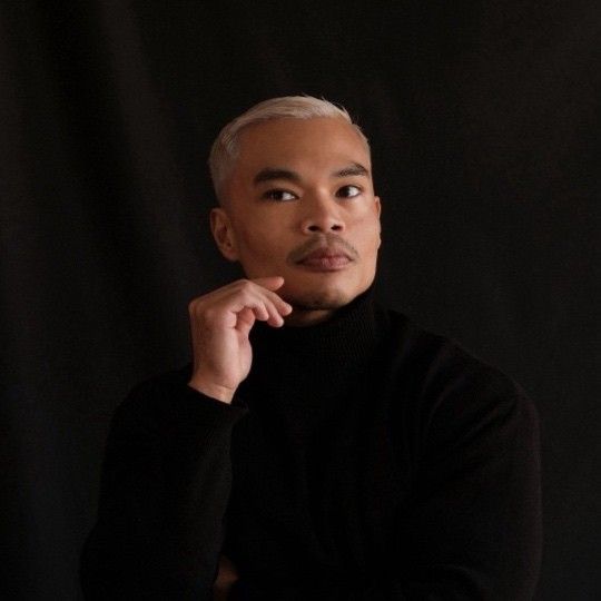 A person with short, light-colored hair and a mustache wearing a black turtleneck poses thoughtfully against a dark background.