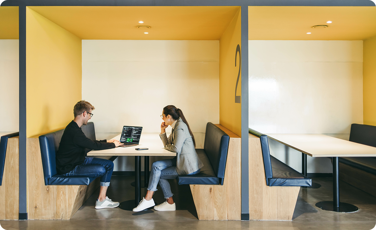Two people sitting across from each other in a booth, with one working on a laptop displaying code. The setting is a modern, open workspace with yellow walls.