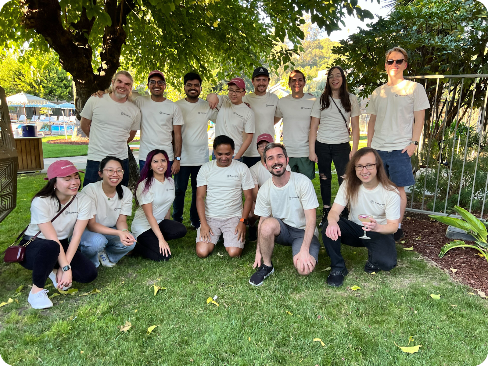 A group of fifteen people wearing matching beige shirts pose under a tree in a grassy area, with smiles and relaxed postures, suggesting a casual outdoor gathering or team event.
