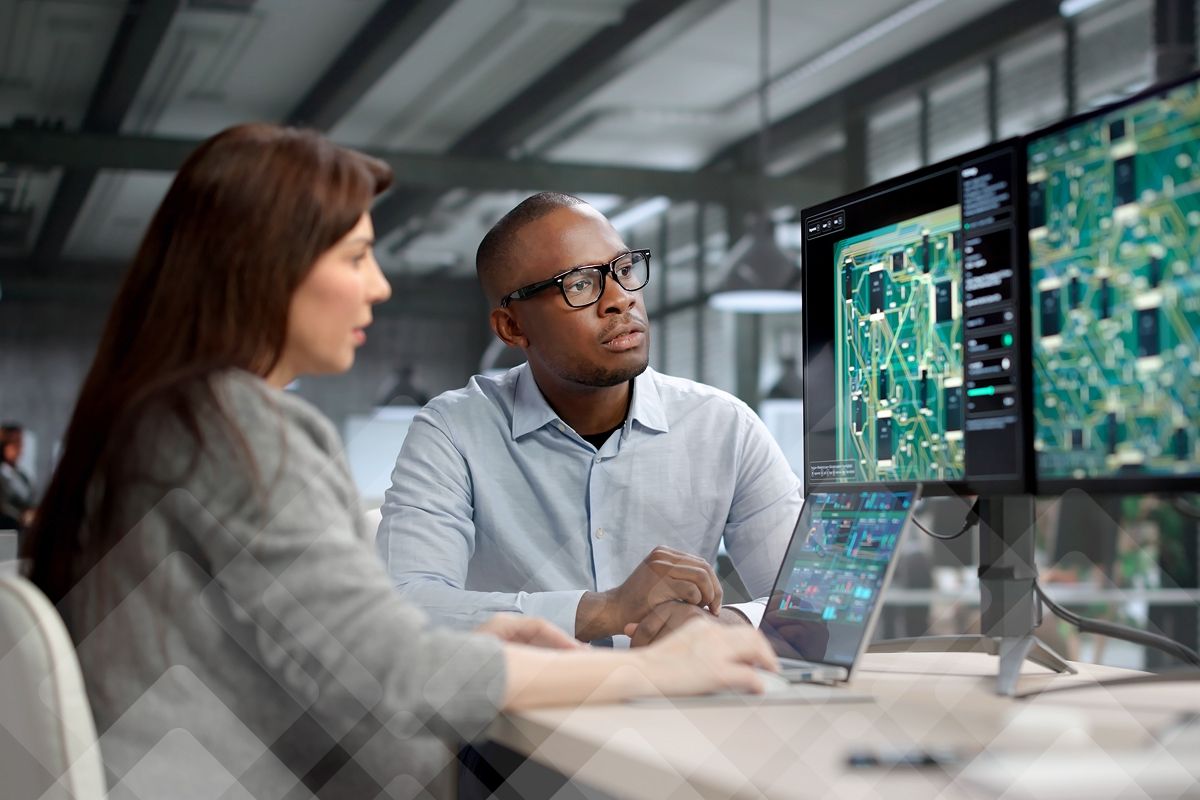 Two colleagues reviewing circuit board designs on dual monitors and a laptop.
