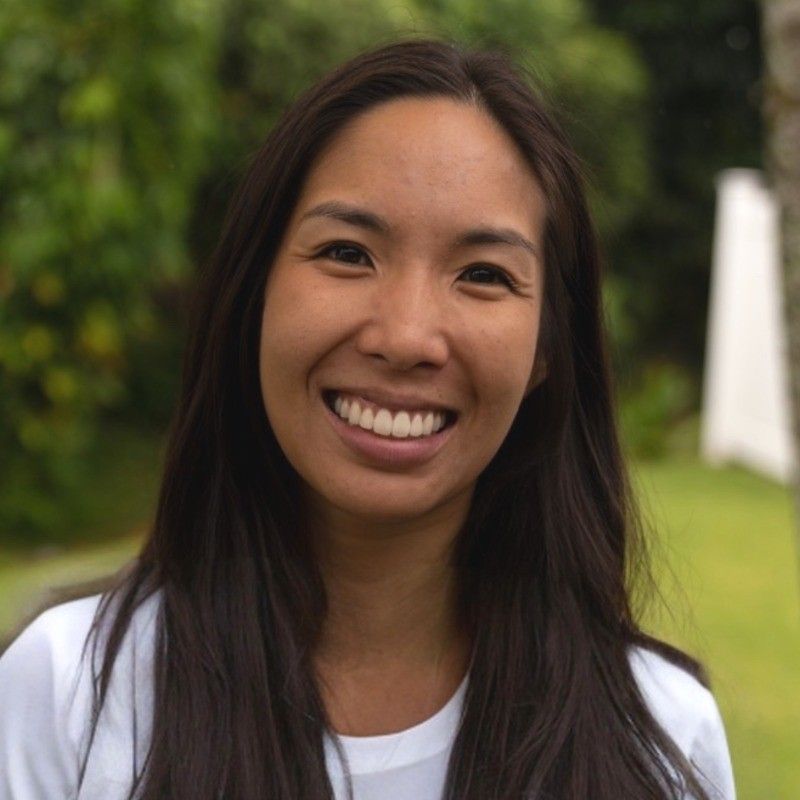 A person with long dark hair smiles warmly, standing in a garden setting with greenery in the background.