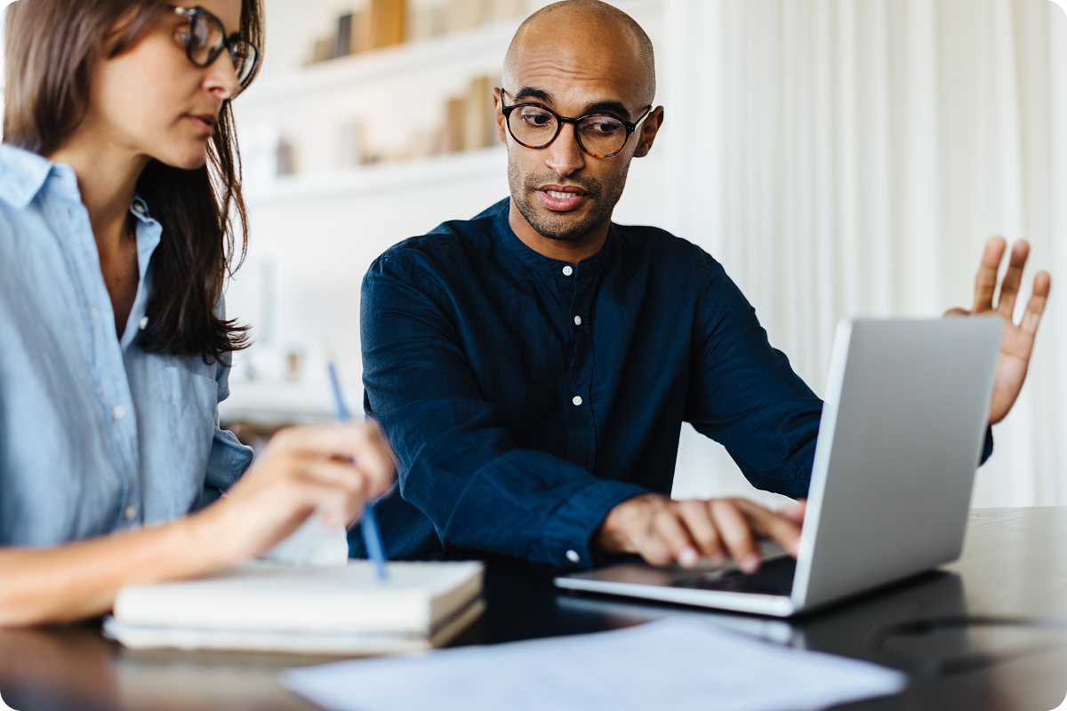 A man and a woman sit at a table, both wearing glasses and casual clothing. The man gestures while looking at a laptop, and the woman writes in a notebook.