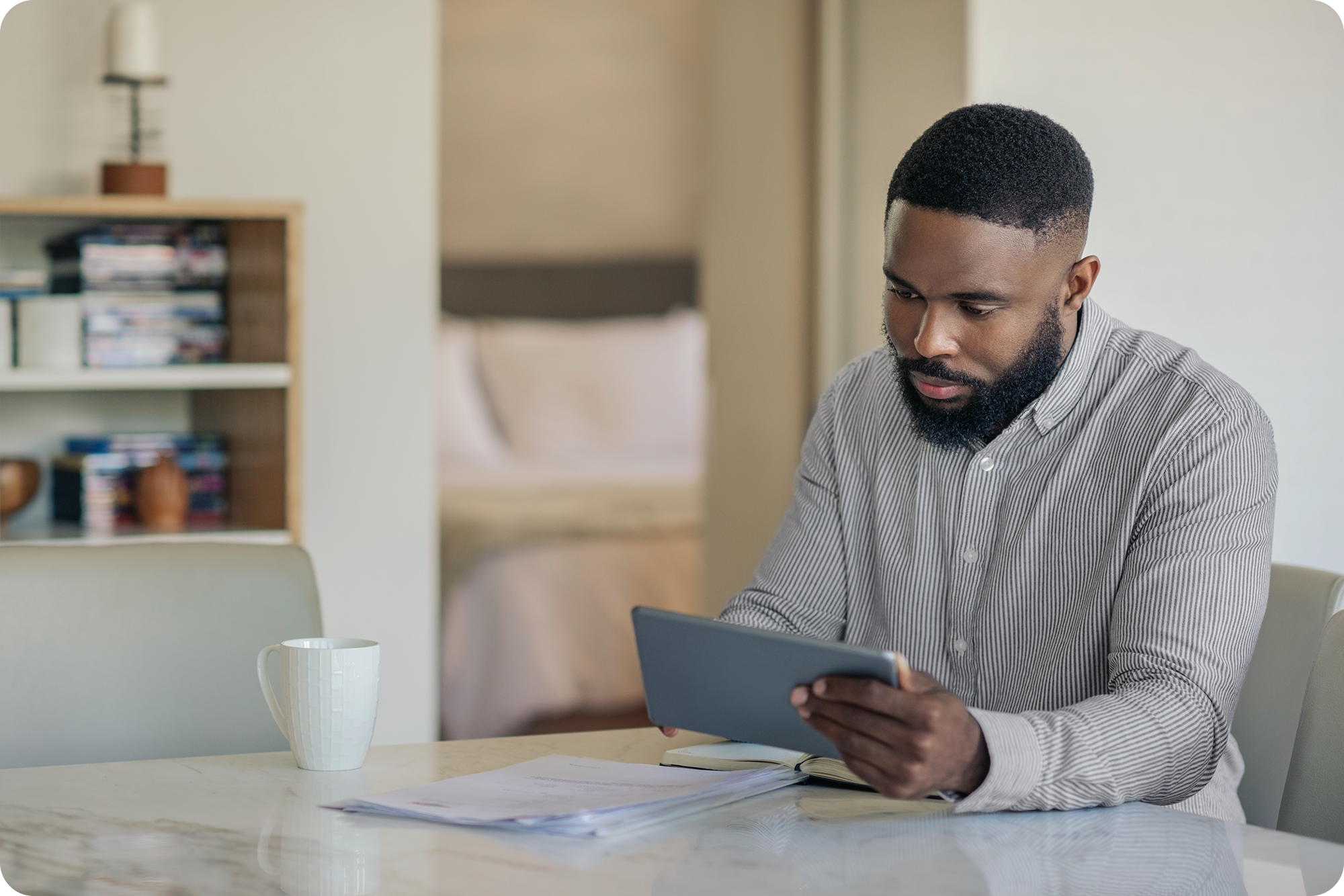 A man with a beard sits at a table reading a tablet, surrounded by papers and a coffee mug, in a modern room with books on a shelf in the background.