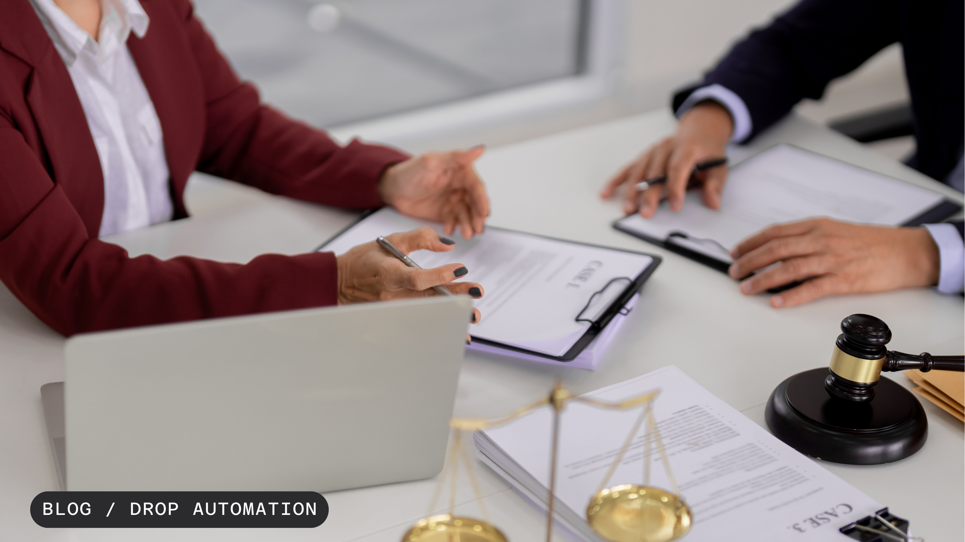 Hands of two people discussing documents at a desk with a laptop, gavel, and scales of justice.