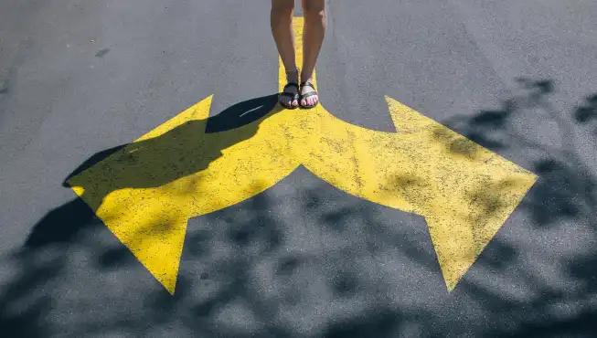 Person standing on a road with large yellow arrows pointing in opposite directions painted on the surface.