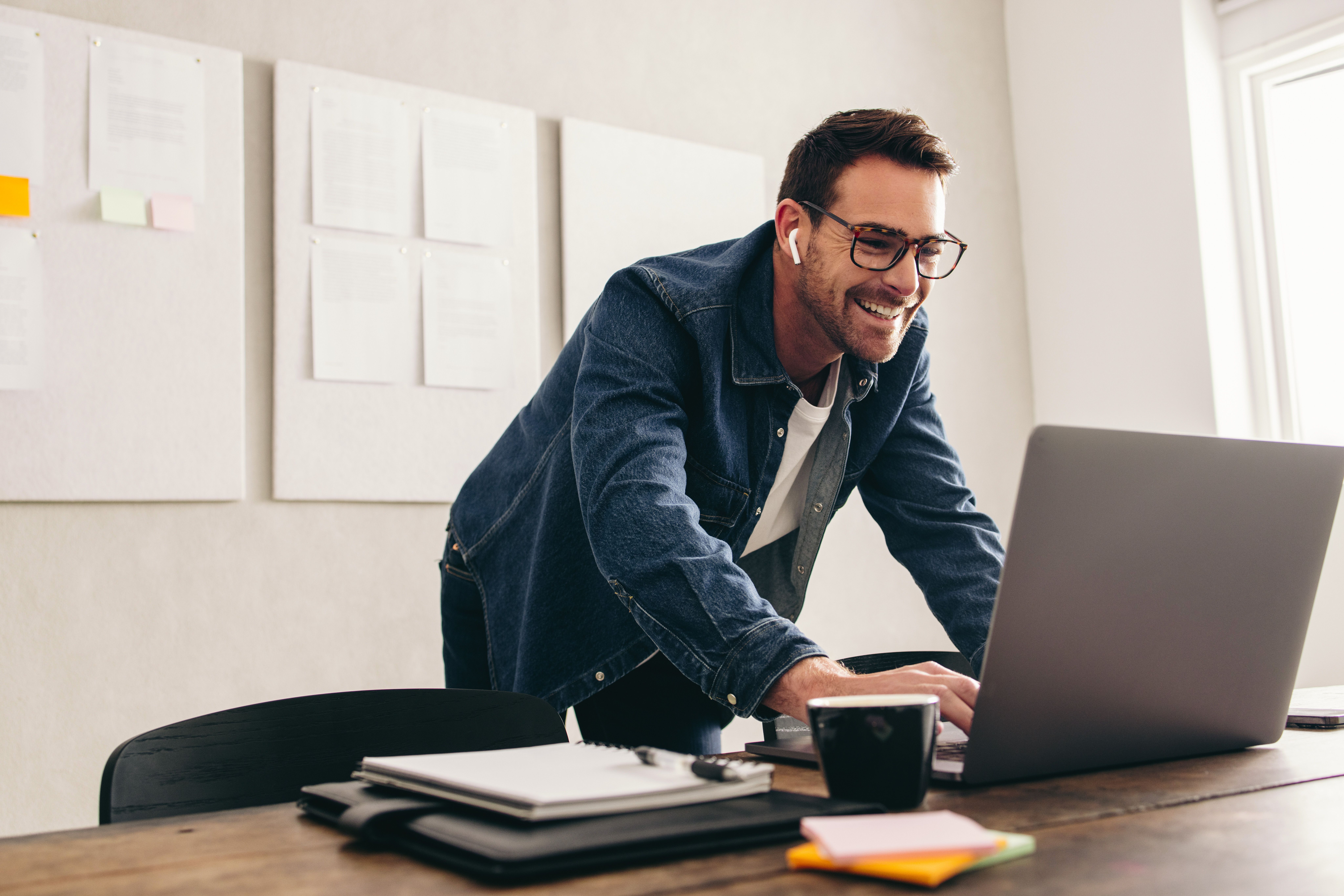 A man wearing glasses and earphones smiles while working on a laptop at a desk covered with notepads and a coffee cup, with whiteboards and papers hanging on the wall behind him.