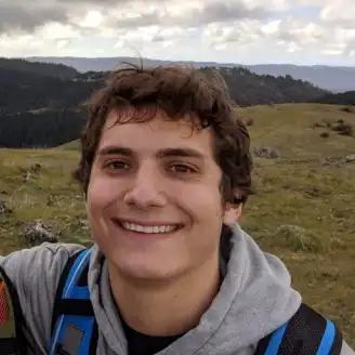 A young man with curly hair and a backpack smiles broadly while standing outdoors on a grassy hillside, with a cloudy sky and distant hills in the background.