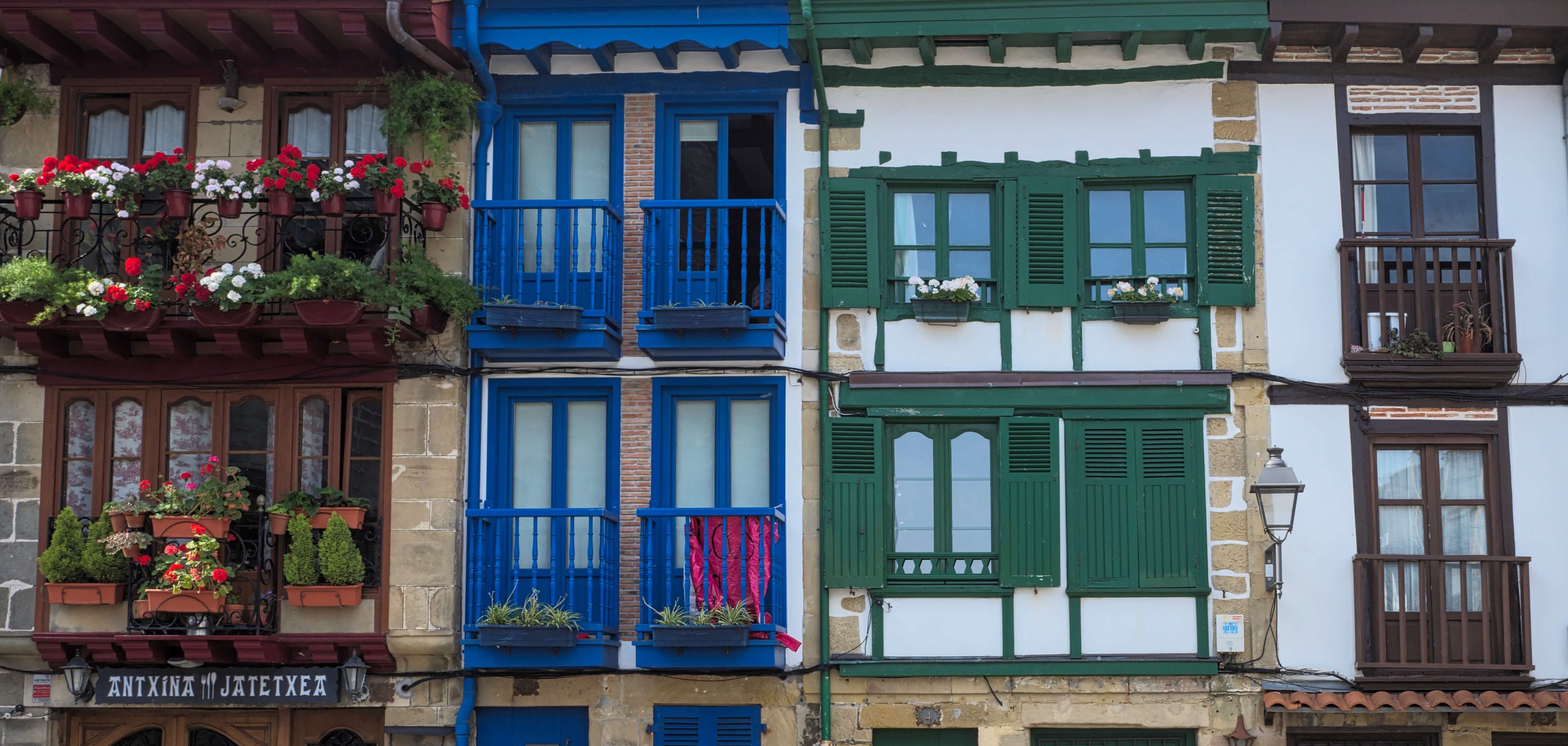 Colorful traditional buildings with vibrant balconies and green shuttered windows, adorned with potted flowers, are shown in a quaint street setting.