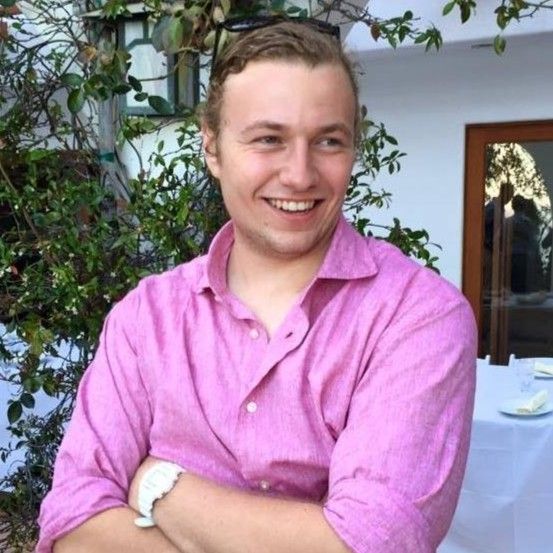 Person in a pink shirt smiling with arms crossed, standing in front of greenery and a building with a window.