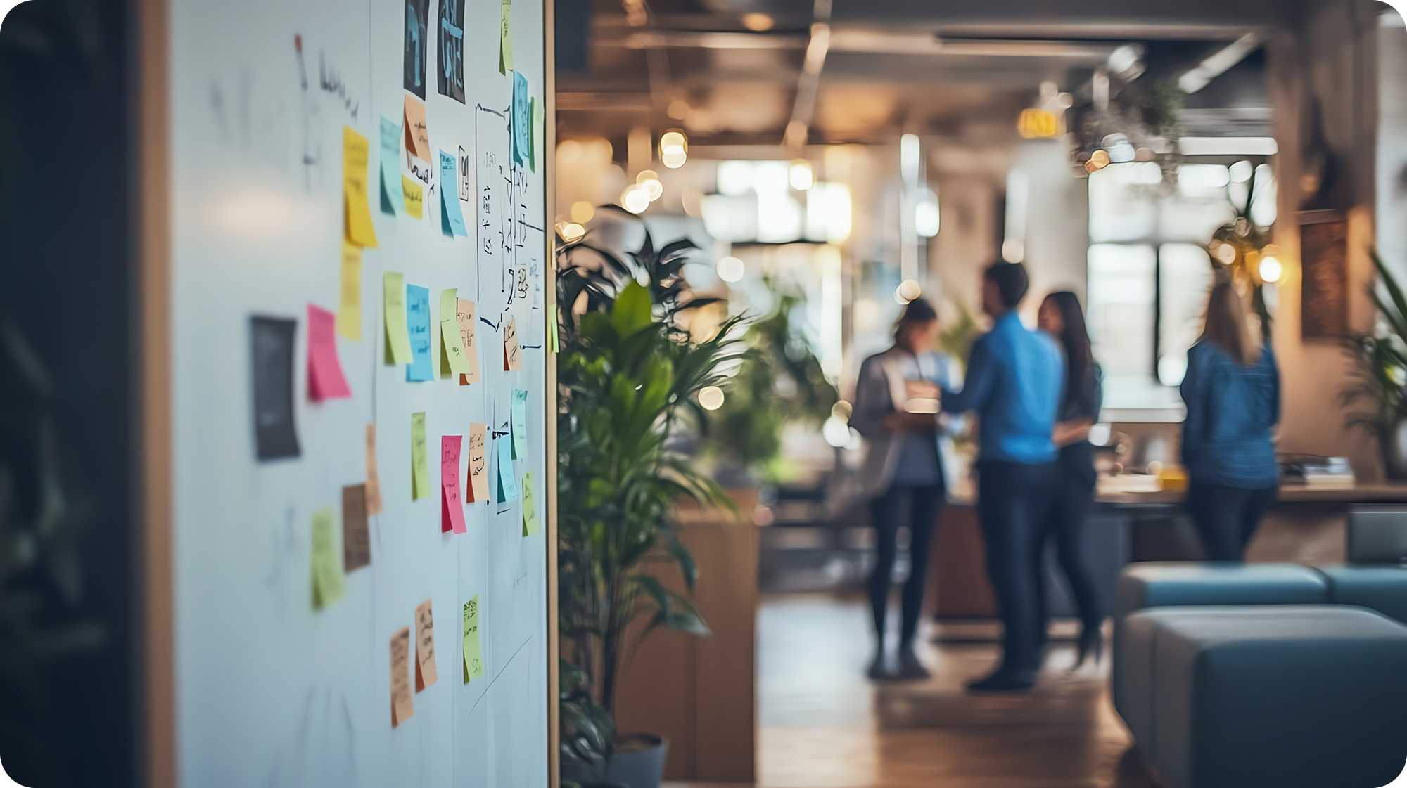 A whiteboard filled with colorful sticky notes is in the foreground, while a group of people engage in conversation in a modern, brightly lit office space in the background. Several plants and soft seating create a relaxed atmosphere.