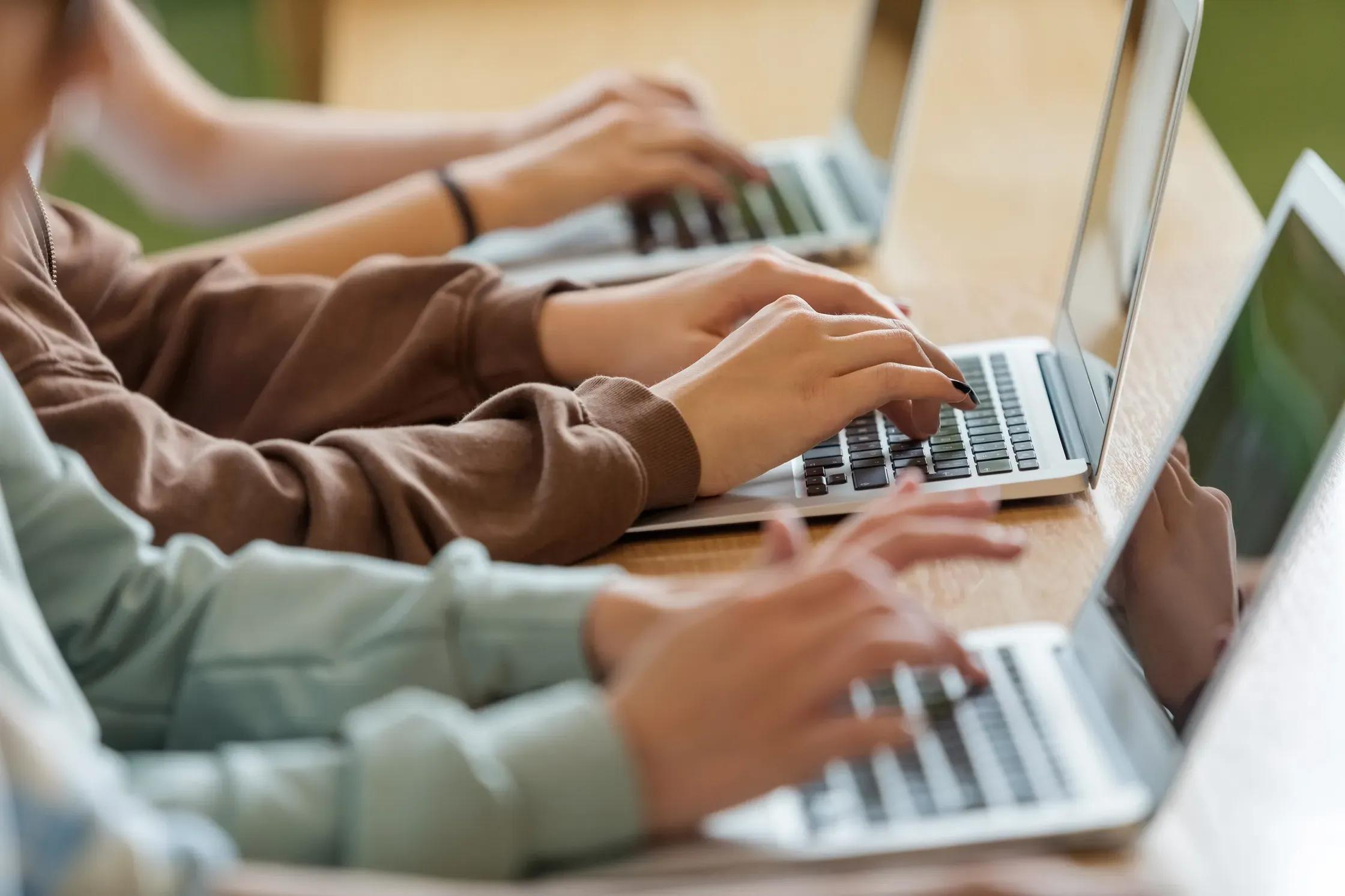 Several people sit side by side typing on laptops, with focus on their hands and keyboards.