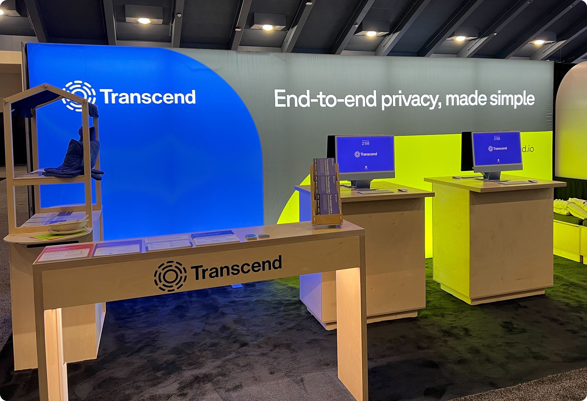 A trade show booth features a bright blue and green color scheme with the Transcend logo and the slogan "End-to-end privacy, made simple." Two monitors are displayed on wooden stands, alongside informational materials.
