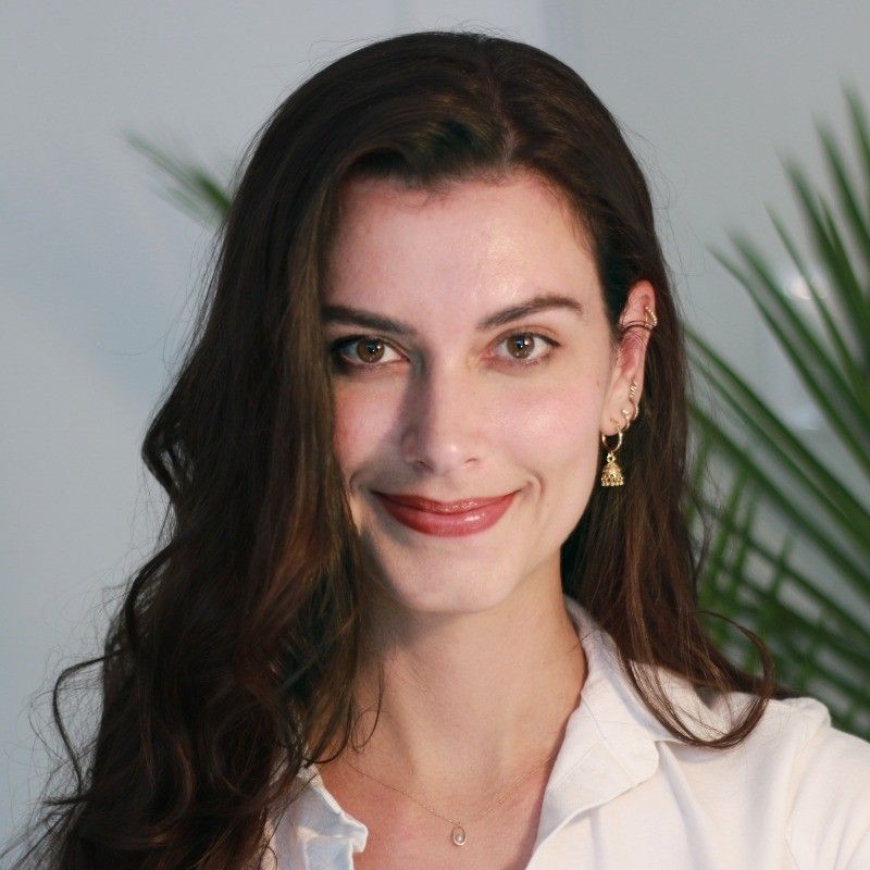 A woman with long brown hair smiles softly, wearing a white blouse and gold earrings, with green plants in the blurred background.