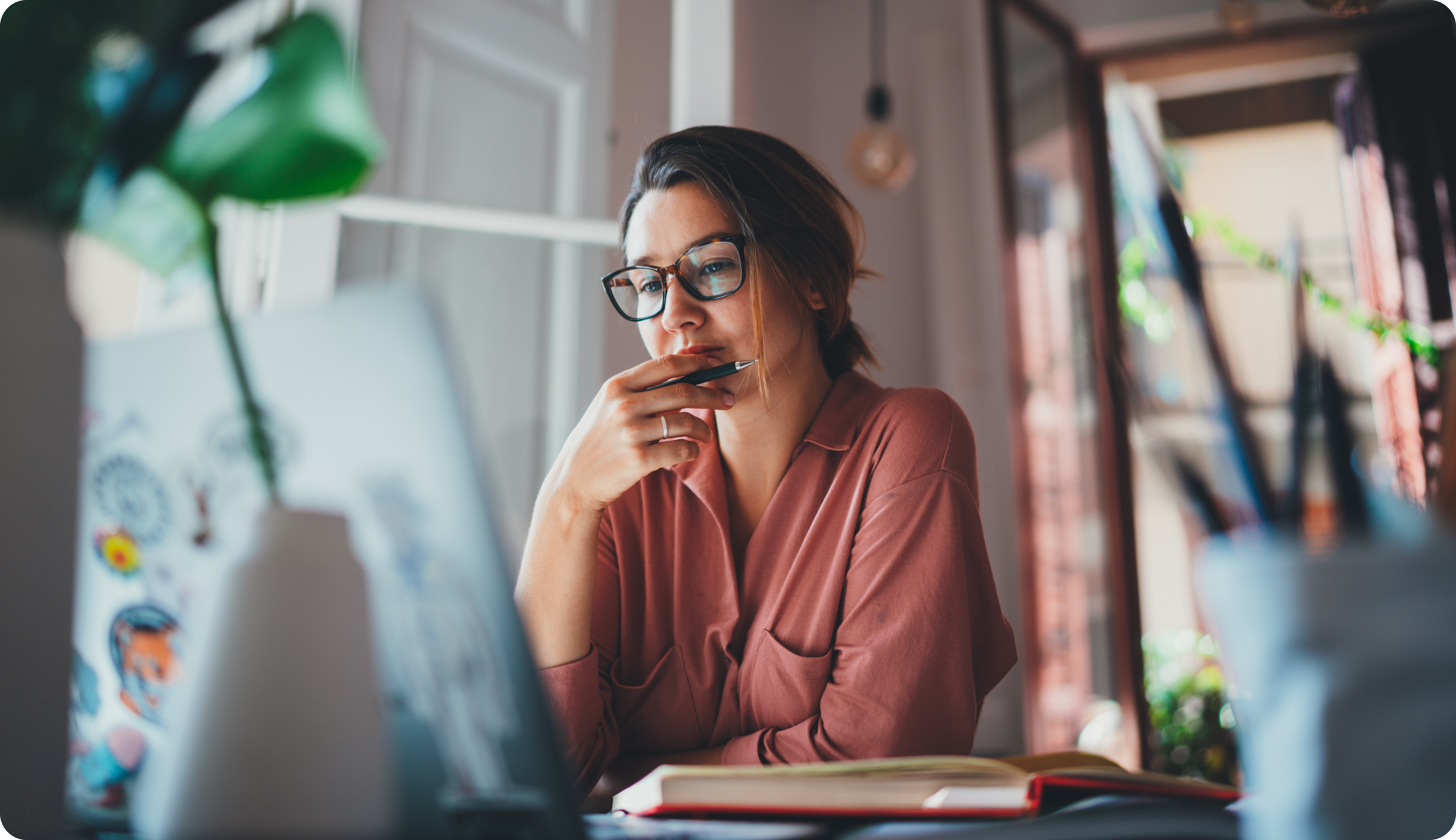 A woman wearing glasses sits at a desk, thoughtfully looking at her laptop with a pen in hand, surrounded by books and colorful decor.