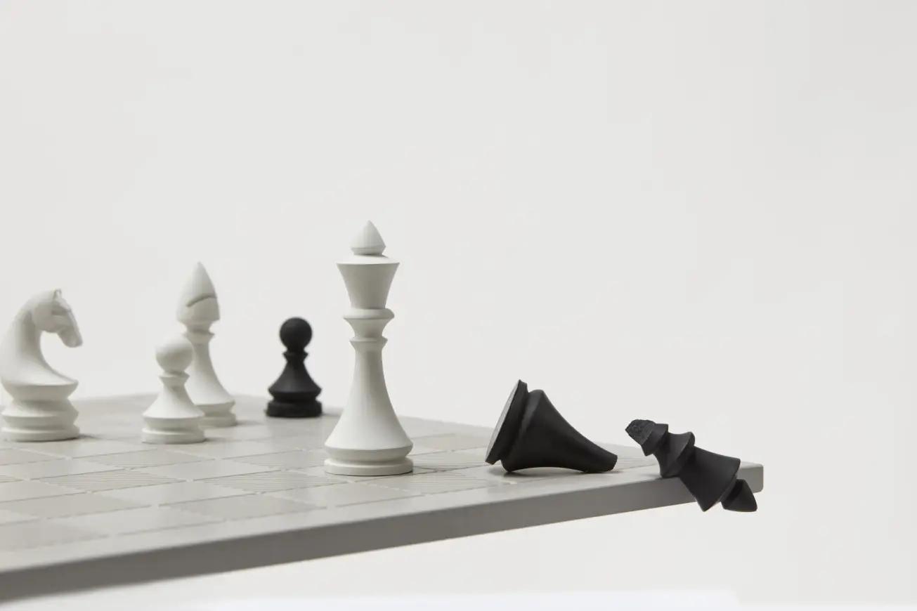 A minimalist chess set features white and black pieces on a board, with a black king falling off the edge, while other pieces remain on the board.