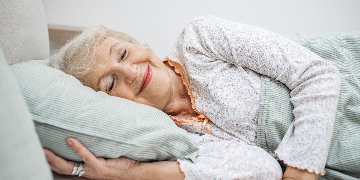 older woman laying in bed holding pillow with one hand while smiling